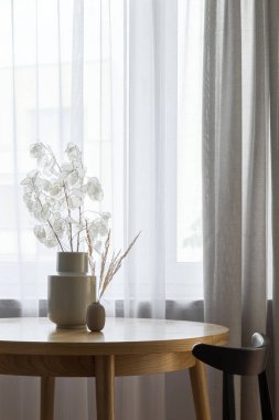Yuvarlak, ahşap yemek masası olan güzel bir oda. Pencerenin yanında beyaz ve bej perdeleri olan doğal bir dekorasyon.