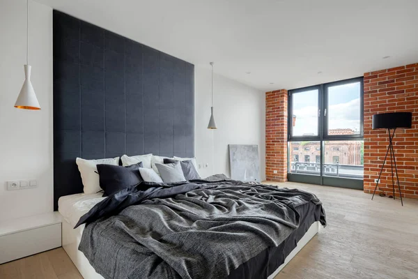 Elegant bedroom with industrial style wall with exposed red brick, big comfortable bed with upholstered wall and big window
