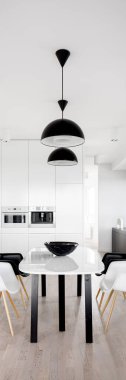 Modern, siyah ve beyaz yemek masasının dikey manzarası. Ahşap zeminli şık bir mutfakta, iki siyah tavan lambası altında sandalyeler.