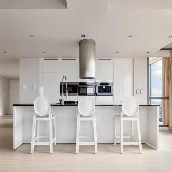 Elegant kitchen with white furniture, modern household appliances and long kitchen island with black countertop and white, stylish chairs