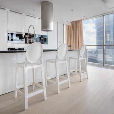 Büyük camları, beyaz dolapları, mutfak adası ve modern beyaz sandalyeleri olan zarif bir mutfak.