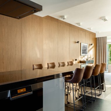 Ahşap duvarlı zarif mutfak ve indüksiyon hobisi, fırın ve sandalyeli yemek masası olan uzun mutfak adası.