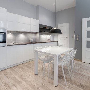 Gri duvarları, ahşap zemini, beyaz dolapları, çekmeceleri ve yeni beyaz sandalyeli modern beyaz yemek masası olan sade ve şık bir mutfak.