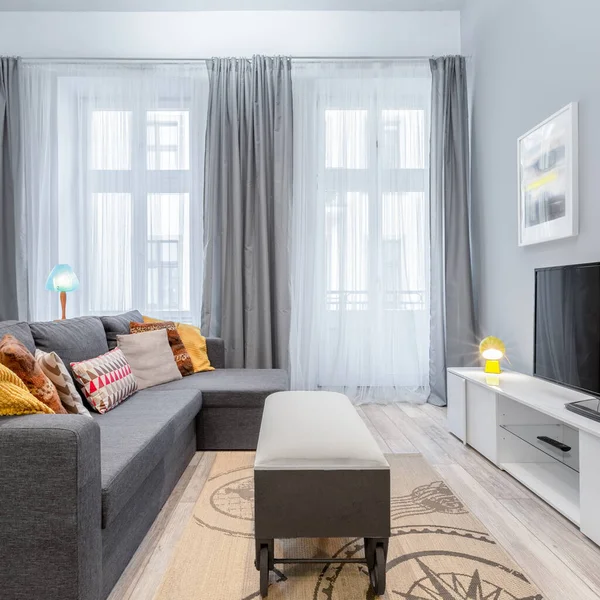 Small living room with grey corner sofa and stylish coffee table on wheels and big windows behind gray and white curtains