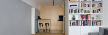 Mutfağı açık, beyaz kitaplığı ve gri duvarında dekoratif betonu olan bir apartman manzarası.
