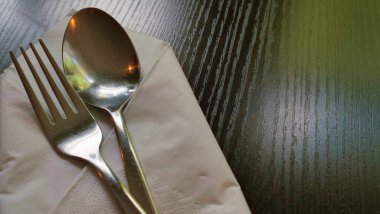 Paslanmaz çelik çatal ve kaşığın düzgünce katlanmış beyaz kağıt peçeteye yerleştirildiği minimalist bir yemek düzeninin yakın çekimi. Çatal bıçaklar görünür tahıl dokusuyla karanlık bir tahta masanın üzerinde duruyor. Sıcak ve davetkar bir atmosfer yaratıyorlar..