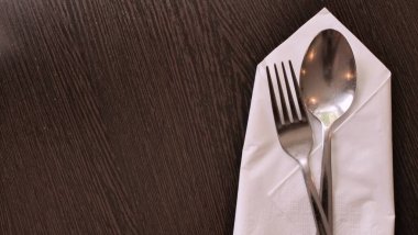 Paslanmaz çelik çatal ve kaşığın düzgünce katlanmış beyaz kağıt peçeteye yerleştirildiği minimalist bir yemek düzeninin yakın çekimi. Çatal bıçaklar görünür tahıl dokusuyla karanlık bir tahta masanın üzerinde duruyor. Sıcak ve davetkar bir atmosfer yaratıyorlar..
