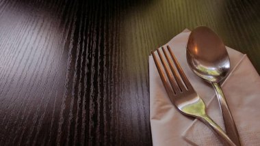 Paslanmaz çelik çatal ve kaşığın düzgünce katlanmış beyaz kağıt peçeteye yerleştirildiği minimalist bir yemek düzeninin yakın çekimi. Çatal bıçaklar görünür tahıl dokusuyla karanlık bir tahta masanın üzerinde duruyor. Sıcak ve davetkar bir atmosfer yaratıyorlar..