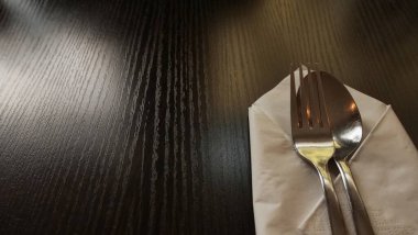 Paslanmaz çelik çatal ve kaşığın düzgünce katlanmış beyaz kağıt peçeteye yerleştirildiği minimalist bir yemek düzeninin yakın çekimi. Çatal bıçaklar görünür tahıl dokusuyla karanlık bir tahta masanın üzerinde duruyor. Sıcak ve davetkar bir atmosfer yaratıyorlar..