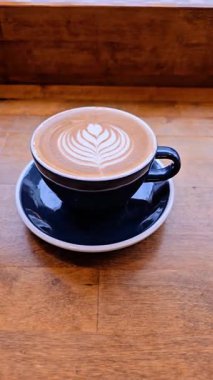 Koyu seramik bir bardakta kapuçinoya zoom yap, dokulu ahşap bir masada, koyu tonlu. Latte sanatı karamsar arkaplana karşı duruyor, sıcak ve davetkar bir kafe atmosferi yaratıyor..