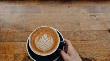 Ahşap masada latte sanatıyla birlikte koyu mavi bir kahve fincanı tutuyor. Konforlu kafe havası, kahveci, hayat tarzı, rahatlama, sabah rutini, latte yakın çekim, estetik içecek, sıcak tonlar, yavaş yaşam. İdeal stok görüntüsü.