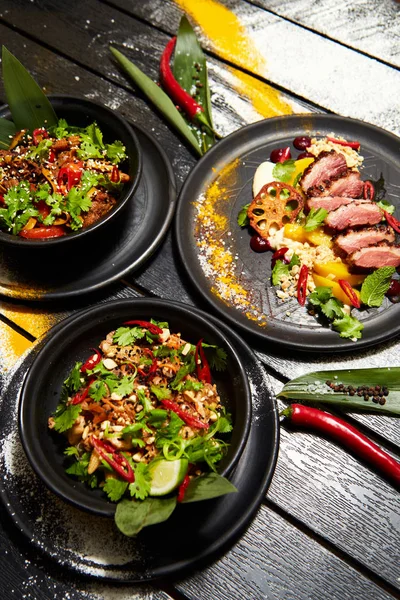 Siyah bir masada Asya yemekleri. Şeftali ile Ördek göğsü, sığır eti ile erişte, tavuk ve karides ile pad tay.