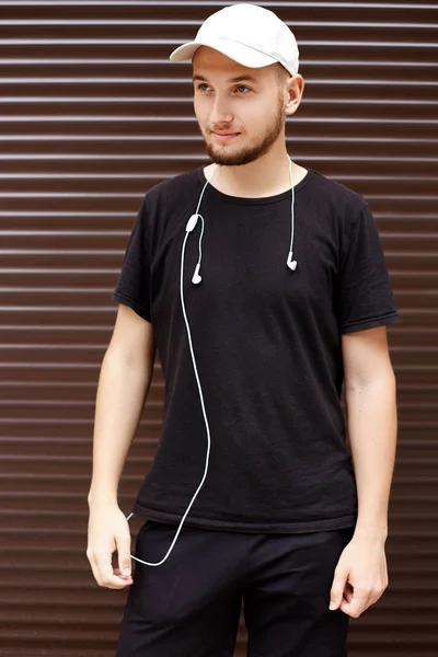 Knappe kerel in een witte Pet en zwart t-shirt met koptelefoon op straat — Stockfoto