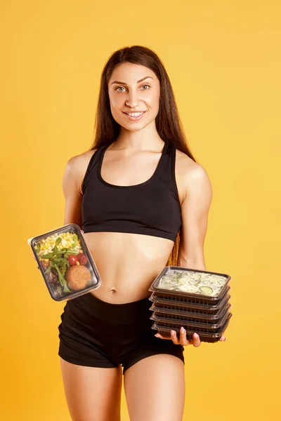 Muchacha atlética atractiva vestida con una camiseta deportiva y pantalones cortos posando en el estudio sobre un fondo amarillo con cajas de comida. Concepto de alimentación saludable, dieta diaria —  Fotos de Stock