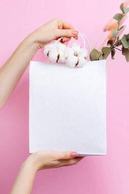 Kadın eli, pembe arka planda çiçeklerle dolu beyaz el çantası tutuyor.