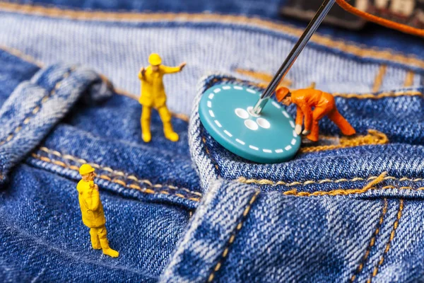 The concept for the fashion industry. Miniature workers sew on buttons big needle to jeans. Close up.