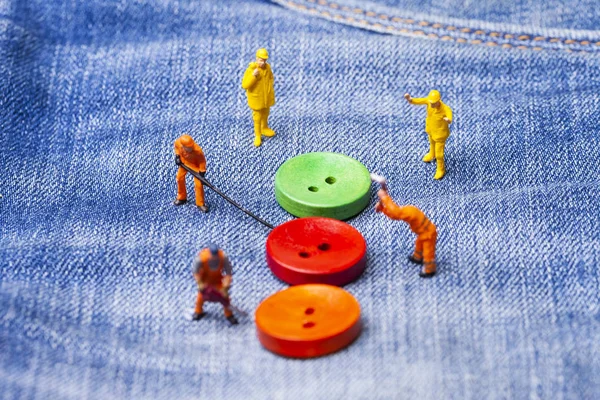 The concept for the fashion industry. Miniature workers sew on buttons big needle to jeans. Close up.