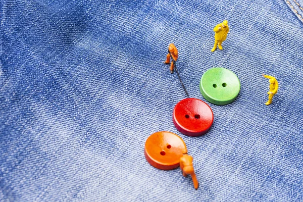 The concept for the fashion industry. Miniature workers sew on buttons big needle to jeans. Close up.