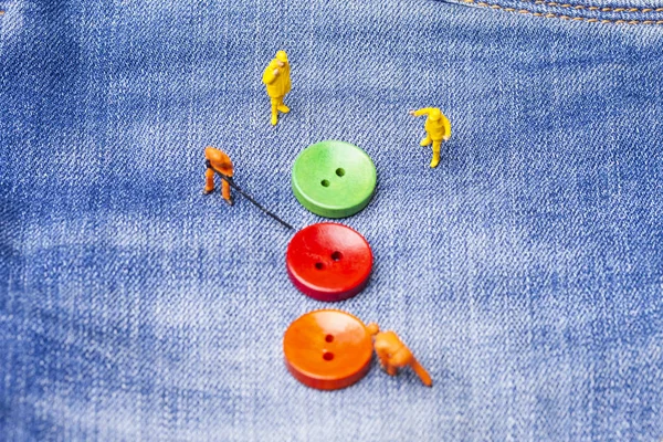 The concept for the fashion industry. Miniature workers sew on buttons big needle to jeans. Close up.