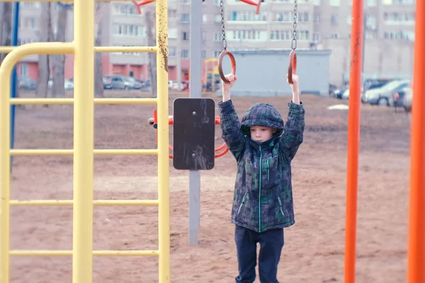 Мальчик качается на гимнастических кольцах, прыгает и улыбается. Детская площадка во дворе городского дома . — стоковое фото