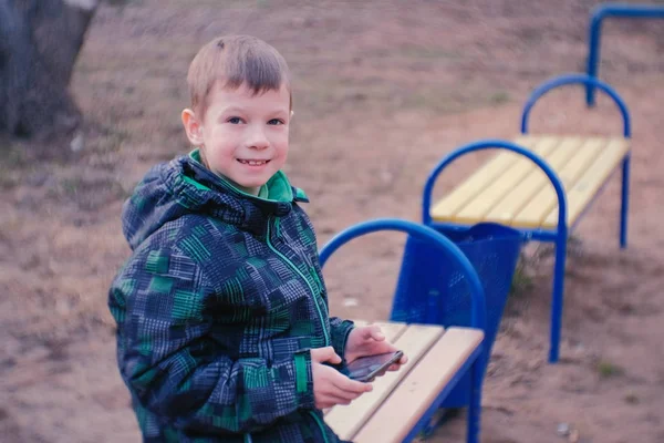 Chico juega un juego en su teléfono móvil sentado en el parque en un banco. Mirando a la cámara y sonriendo . — Foto de Stock
