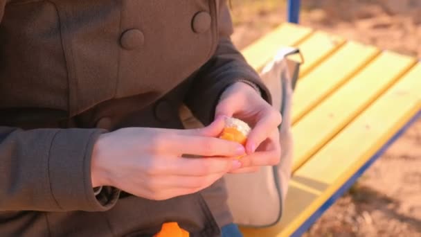 Молода брюнетка в сонцезахисних окулярах пілінгує мандарин, сидячи на лавці в парку. Крупним планом руки . — стокове відео