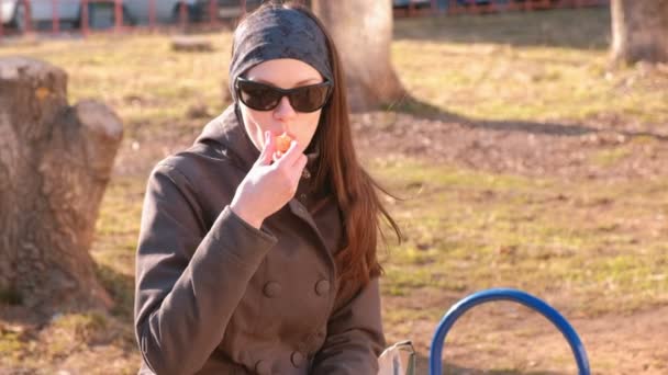 サングラスの若いブルネットの女性は タンジェリン 公園のベンチに座って食べる — ストック動画