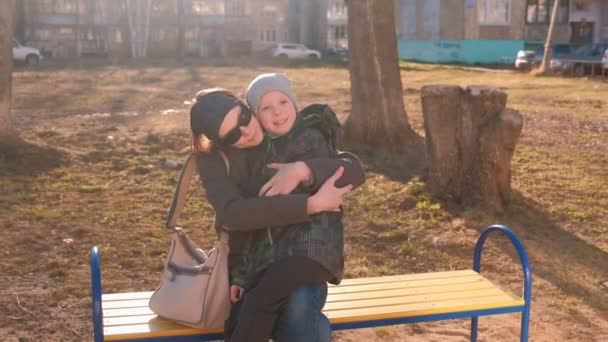 Mom kisses and hugs her son sitting on a bench in the courtyard of a multi-storey building. — Stock Video