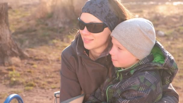 Mamá e hijo miran a su alrededor y hablan sentados en un banco en el parque en la primavera . — Vídeos de Stock