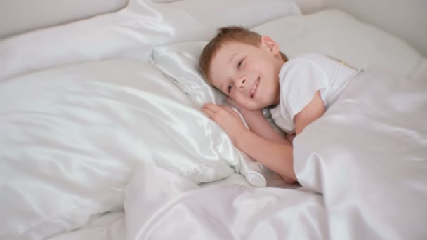 Семилетний мальчик только что проснулся и смеется, лежа в постели. . — стоковое видео