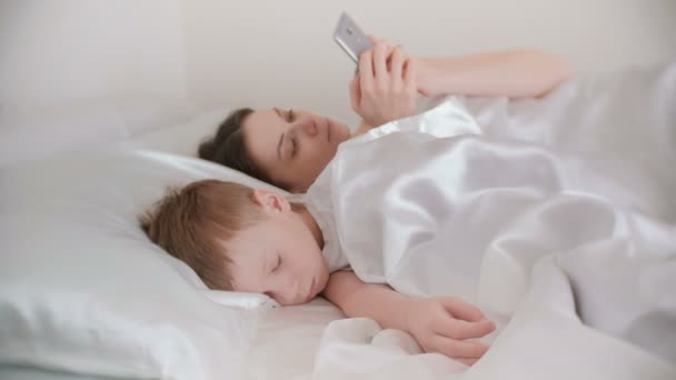 Moeder wakker en surfen op internet in haar mobiele telefoon terwijl haar zoon nog steeds in slaap is. — Stockvideo