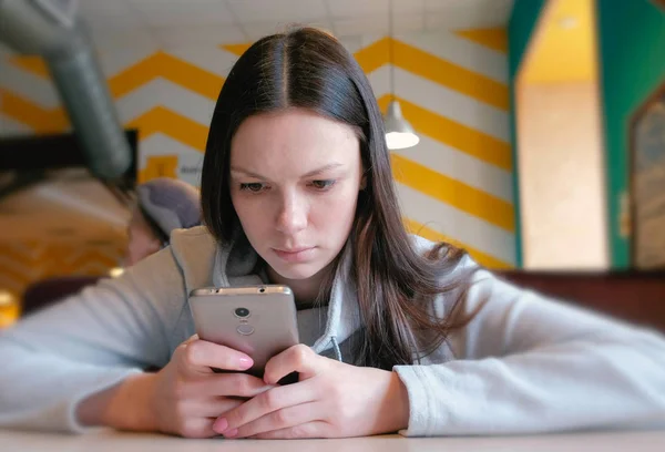 Joven mujer seria leyendo algo en su teléfono móvil sentado en la cafetería . — Foto de Stock