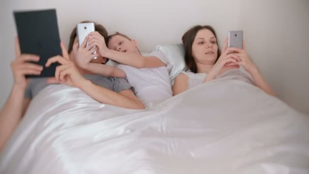 Rodzina jest za pomocą urządzeń mobilnych telefonów i tabletu leżąc w łóżku rano. — Wideo stockowe