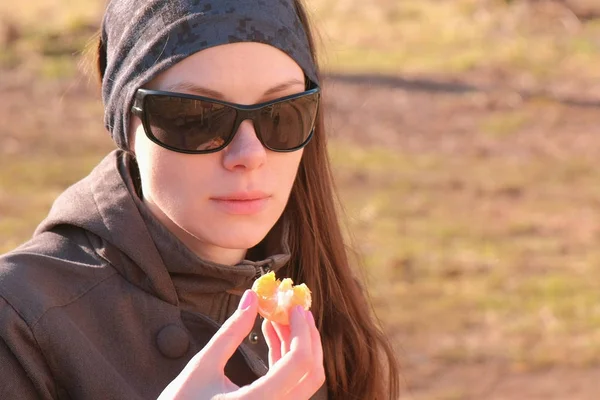 Ung brunett kvinna i solglasögon äter tangerine sitter på bänken i parken. Närbild ansikte. — Stockfoto