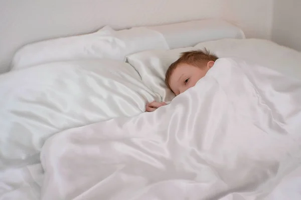 Семилетний мальчик просыпается в своей постели под одеялом — стоковое фото