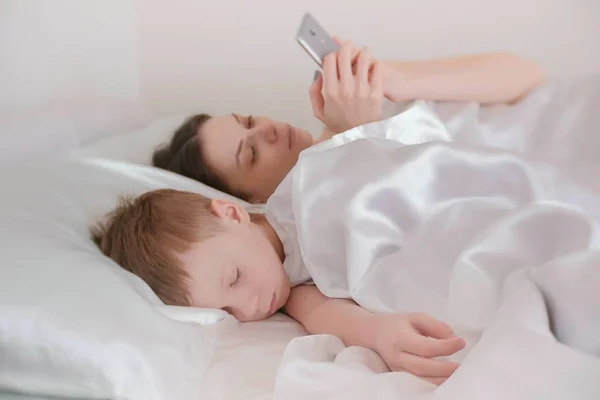 Мама проснулась и просматривает интернет в своем мобильном телефоне. Её сын всё ещё спит. . — стоковое фото