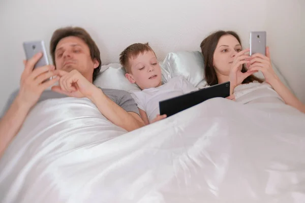 La familia está utilizando teléfonos móviles y tabletas que yacen en la cama por la mañana . — Foto de Stock