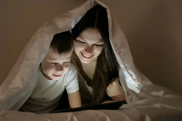 Mujer joven mamá y su hijo viendo una película juntos en la tableta debajo de la manta . — Foto de Stock