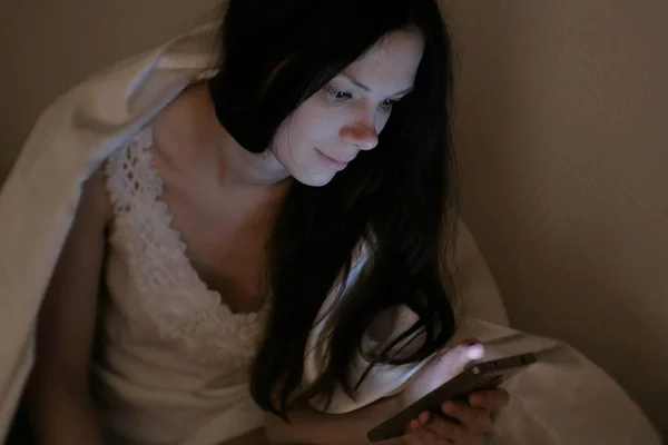 Jovem bela mulher morena olha para as redes sociais em seu telefone celular na cama antes de dormir e sorri . — Fotografia de Stock