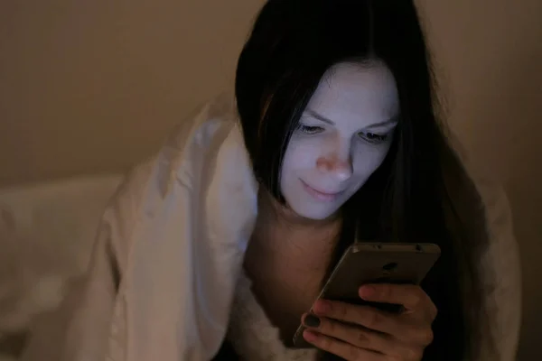 Brunette fiatal gyönyörű nő néz szociális hálózatok neki mobil telefon az ágyban alszik, és mosolyog. — Stock Fotó