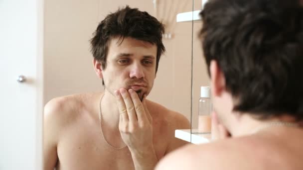 Hombre cansado que acaba de despertar mirando su reflejo en el espejo y se frota la barba de la mano — Vídeo de stock