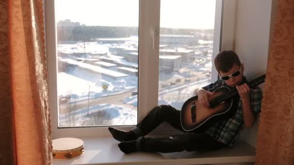 Tocar un instrumento musical. Niño en gafas de sol toca la guitarra sentado en el alféizar . — Vídeos de Stock