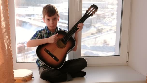 A tocar um instrumento musical. Menino toca guitarra e cantando sentado no peitoril da janela . — Vídeo de Stock
