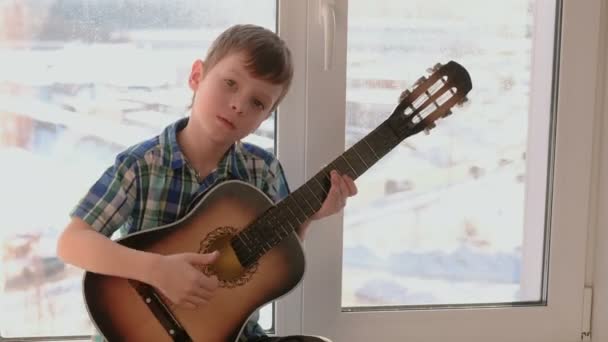 Het spelen van een muziekinstrument. Jongen speelt de gitaar en het zingen zittend op de vensterbank. — Stockvideo