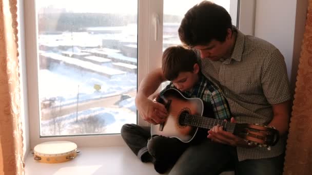 Ein Musikinstrument zu spielen. Papa bringt seinem Sohn das Gitarrespielen bei, sitzt auf der Fensterbank. — Stockvideo