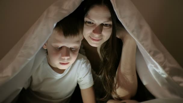 Mladá žena Maminka a její syn společně sledovat zajímavý film na tabletu pod dekou. — Stock video