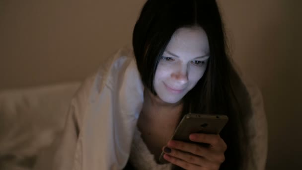 Giovane bella donna bruna guarda i social network nel suo telefono cellulare a letto prima di dormire e sorride . — Video Stock