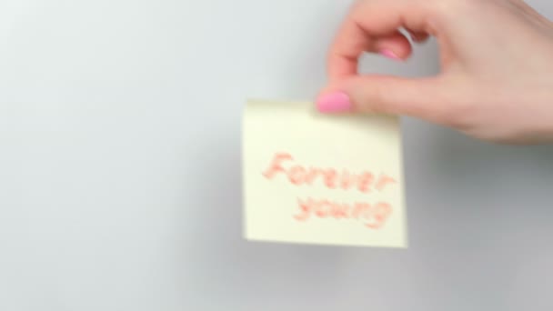 Närbild kvinnans händer stick gul dekal ark med ord för evigt unga. — Stockvideo