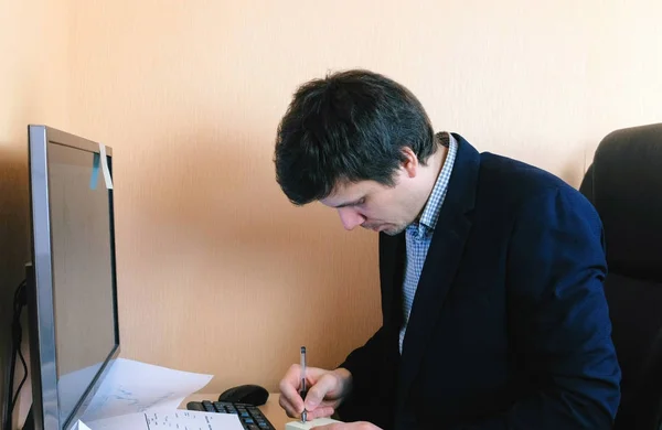 Man aan het werk op de computer. Hij schrijft iets op papier van de sticker. — Stockfoto