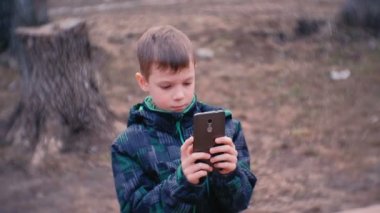 Çocuk cep telefonuna parkta bankta oturmuş bir video izliyor.